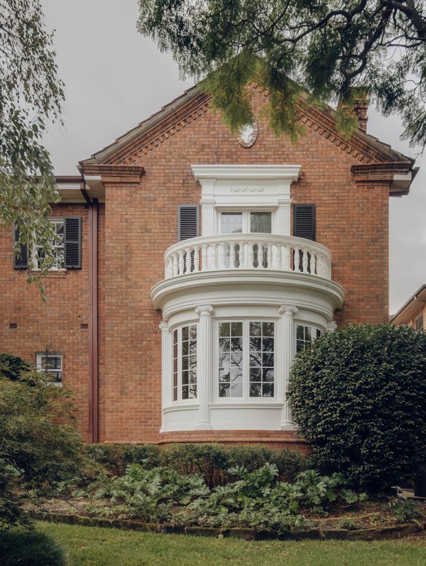 Front exterior view of a grand two-storey red-brick residence with a prominent curved projecting bay window, ornate white classical trim, balustraded balcony, dark shutters, and a steep decorative gabled roof. Mature landscaping frames the facade.