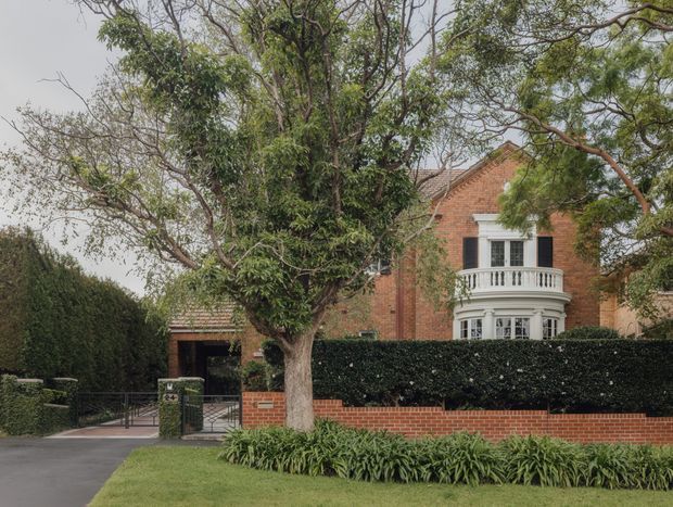 A street-facing view of a substantial red-brick suburban residence, partly screened by mature trees and manicured hedges. The home has a pitched tiled roof, white-painted bay window with a decorative curved balcony, and a low red-brick boundary wall. A paved driveway and gated entry are visible to the left, with established landscaping in the foreground.
