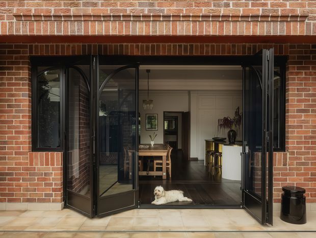 Exterior view of a brick-clad residential entrance with large black-framed bi-fold doors opened onto a tiled threshold. The opening reveals a dark timber-floored interior with a dining table and chairs, framed artwork, a kitchen or bar area, and a small dog lying at the doorway. The image highlights the connection between the outdoor paving and the indoor living space.