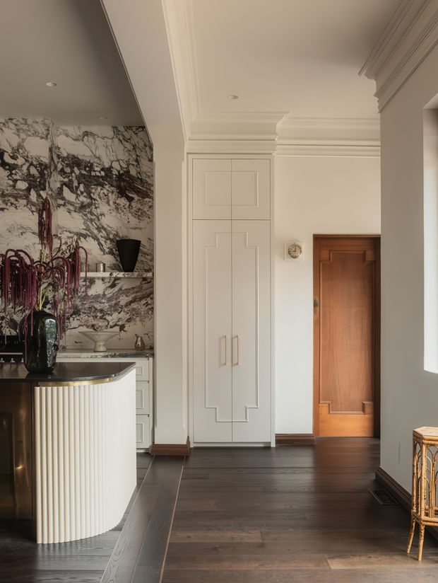 A refined residential interior showing a transitional hallway and kitchen threshold. The space features dark timber flooring, warm white walls with elaborate layered crown moulding and panelled cabinetry, a richly grained timber door, a curved fluted kitchen island with a dark stone benchtop, and a dramatic marble-patterned feature wall. The overall composition is warm, elegant, and highly detailed.