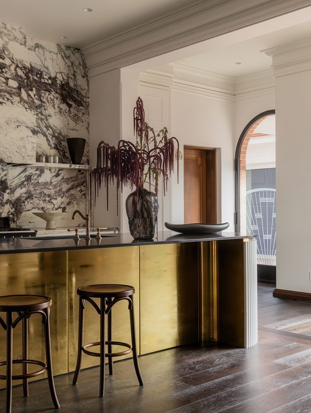 A refined interior bar or kitchen counter with a polished brass-gold front, dark stone benchtop, and two dark timber or metal stools. Dramatic veined marble cladding forms the backsplash and side wall, while ornate ceiling cornices, wall panelling, and an arched opening create a formal period character. A sculptural vase with cascading foliage and dark decorative objects add a luxurious, artful atmosphere.