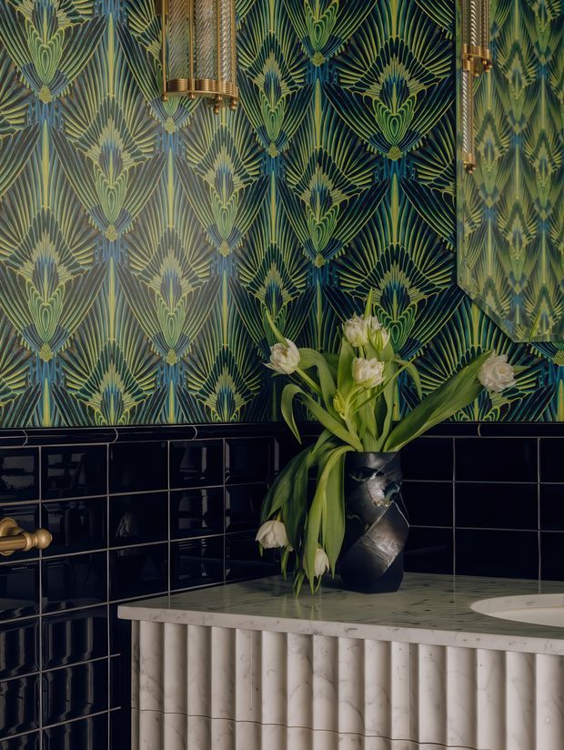 A richly detailed bathroom vanity vignette featuring glossy dark navy wall tiles, an ornate botanical patterned wallcovering, and a pale stone vanity with vertical fluted detailing. A sculptural dark vase with white flowers sits on the counter, while brass wall-mounted tapware and mirror fittings add a warm metallic accent. The overall composition is dramatic, luxurious, and Art Deco-inspired.
