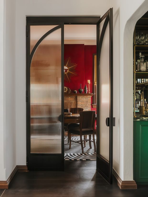A view through black-framed fluted-glass double doors into a richly styled dining room. The room features deep red walls, a brick fireplace with mantel decor, a dark wood dining table and chairs, and a bold black-and-white patterned rug. A partially visible green built-in bar or cabinet with glassware appears at right. The composition has strong Art Deco-inspired geometry, dramatic colour contrast, and polished dark timber flooring.