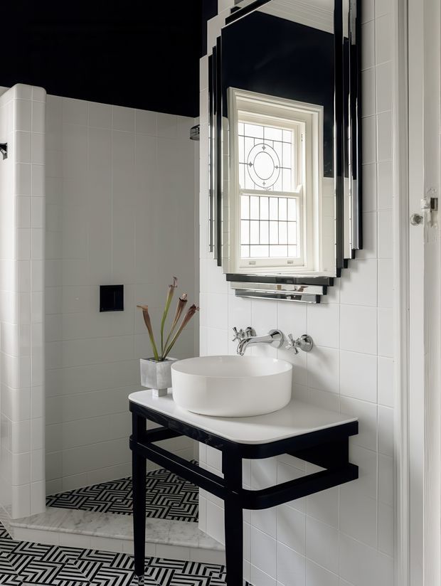 A high-contrast black-and-white bathroom featuring a white vessel basin on a black open-frame vanity, wall-mounted chrome tapware, white tiled walls, a geometric black-and-white tiled floor, and a decorative leaded-glass window framed by a bold black mirror. A shower area is visible to the left.