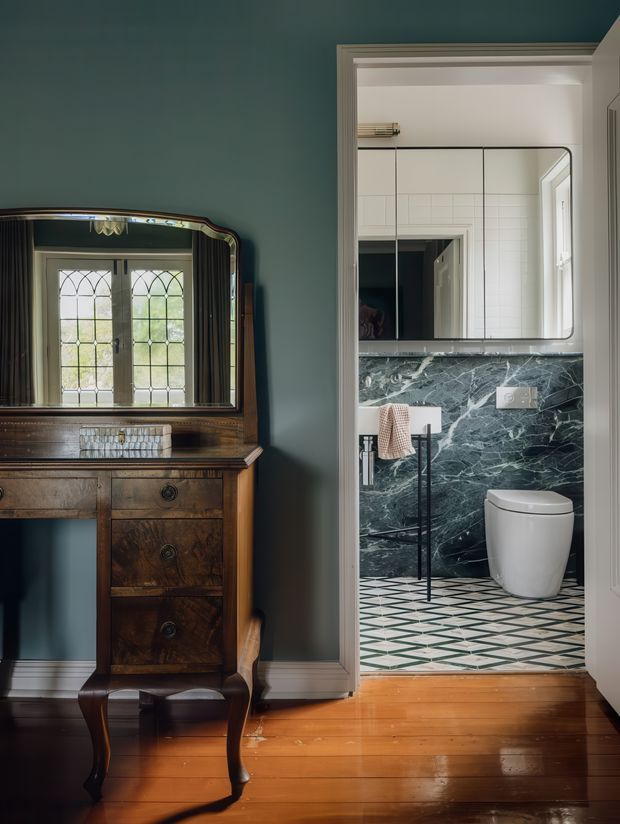 A refined interior view from a dark teal hallway or landing into a compact bathroom. In the foreground is an antique dark timber dressing table with an ornate framed mirror, positioned against the teal wall. The bathroom features white wall tiles, a dark veined stone wall finish, a geometric patterned tiled floor, a wall-mounted black towel rail, white toilet, and a large rounded-corner mirror. Timber flooring and traditional trim frame the doorway, creating a strong contrast between the heritage-style hall and the contemporary bathroom fit-out.