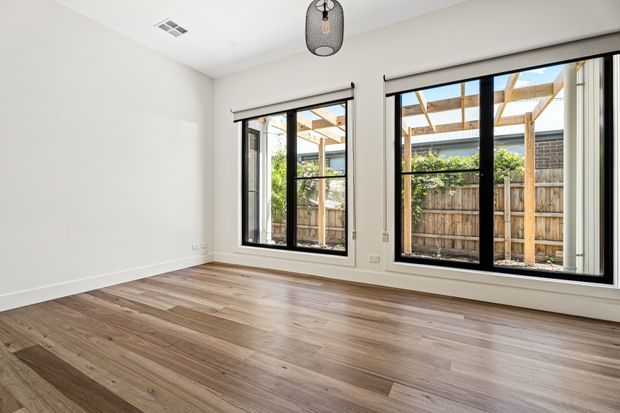 Bright, unfurnished interior room with white walls, wide-plank timber flooring, black-framed windows and a glazed sliding door opening toward a fenced outdoor area with a timber pergola. A simple pendant light and ceiling vent are visible.