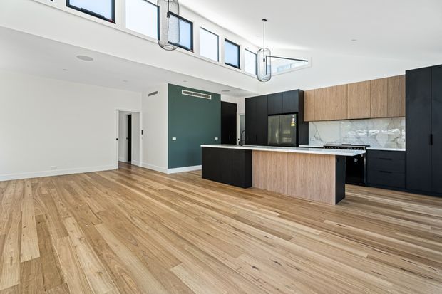 Bright open-plan kitchen and living area with a double-height ceiling and clerestory windows. The kitchen features a large dark island, integrated dark cabinetry, warm timber cabinetry, a marble-look splashback, and contemporary pendant lighting. Wide-plank timber flooring extends across the open space, with a hallway doorway visible at left.