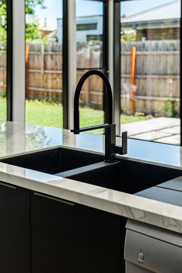 Close-up of a contemporary kitchen sink area featuring a large black double-bowl undermount sink, matte black gooseneck mixer tap, dark cabinetry with slim metal pulls, and a white marble-look benchtop. Full-height black-framed glazing overlooks a fenced backyard and patio.