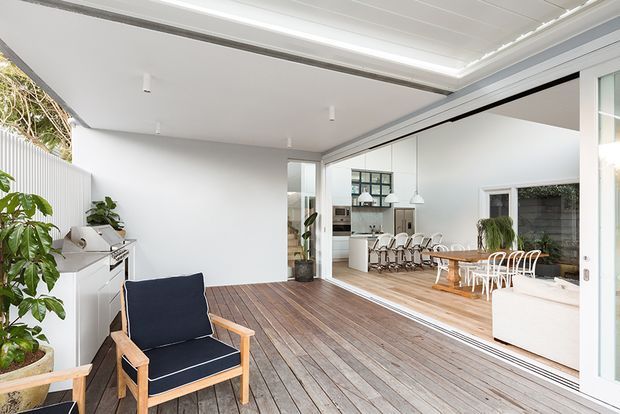 Bright contemporary indoor-outdoor living space with a timber deck opening through wide sliding glass doors into an open-plan dining and kitchen area. The space features white walls and ceilings, light timber flooring, a long dining table with white chairs, built-in cabinetry, potted greenery, and a navy upholstered lounge chair on the deck.