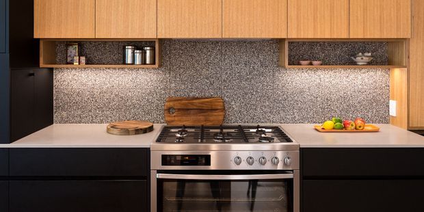 A compact contemporary kitchen with warm timber overhead cabinetry, matte dark lower cabinets, pale benchtops, and a full-height speckled stone or terrazzo-look splashback. Open display shelves, a freestanding stainless-steel gas range, under-cabinet lighting, and minimal countertop accessories create a refined, functional composition.