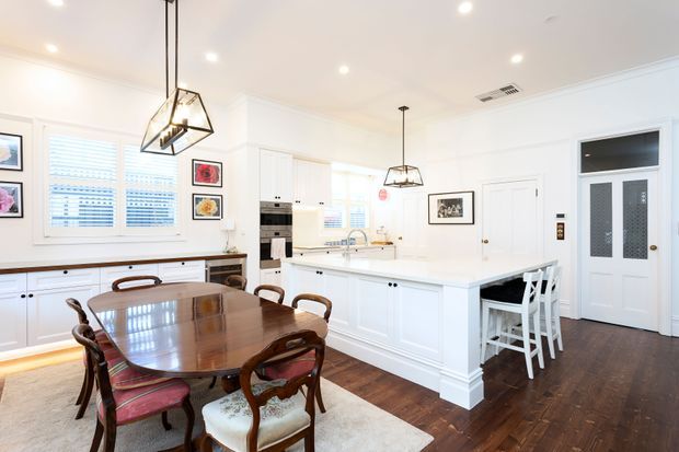 Bright, spacious open-plan kitchen and dining area with white Shaker-style cabinetry, a large island with seating, dark timber flooring, and a polished wooden dining table with upholstered chairs. Recessed ceiling lights and geometric pendant fixtures provide layered lighting, while framed artwork, plantation shutters, and traditional trim add refined character.