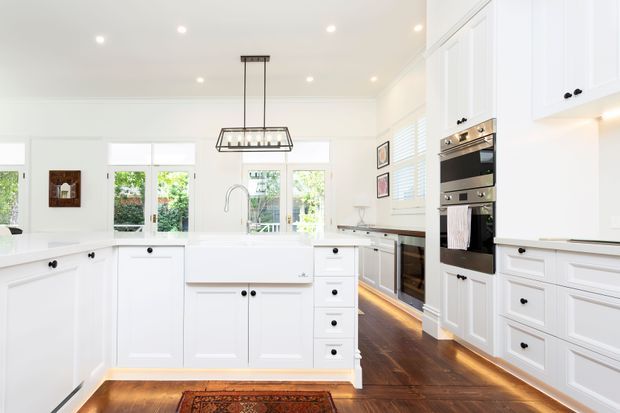 Bright, spacious white kitchen with extensive Shaker-style cabinetry, a central island with undermount sink and gooseneck tap, integrated stainless-steel ovens, dark timber flooring, and a rectangular pendant light. Large windows and plantation shutters bring in abundant natural light, while dark hardware and a patterned rug add contrast.
