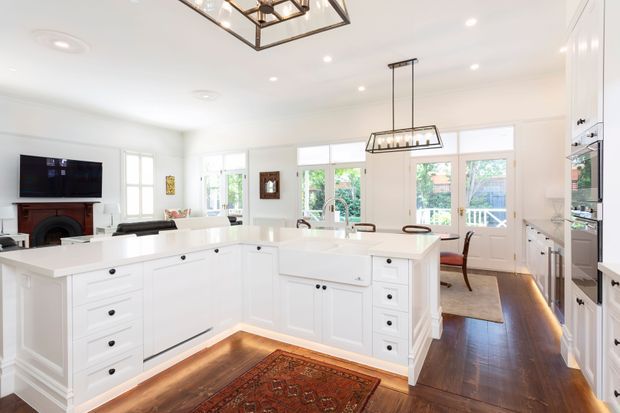 Bright, spacious white kitchen opening into a living and dining area. The room features extensive white shaker-style cabinetry, a large central island with a white benchtop, dark timber flooring, brass-toned hardware, and several geometric pendant lights. French doors and windows provide abundant natural light, while a fireplace and television are visible in the adjoining living area.