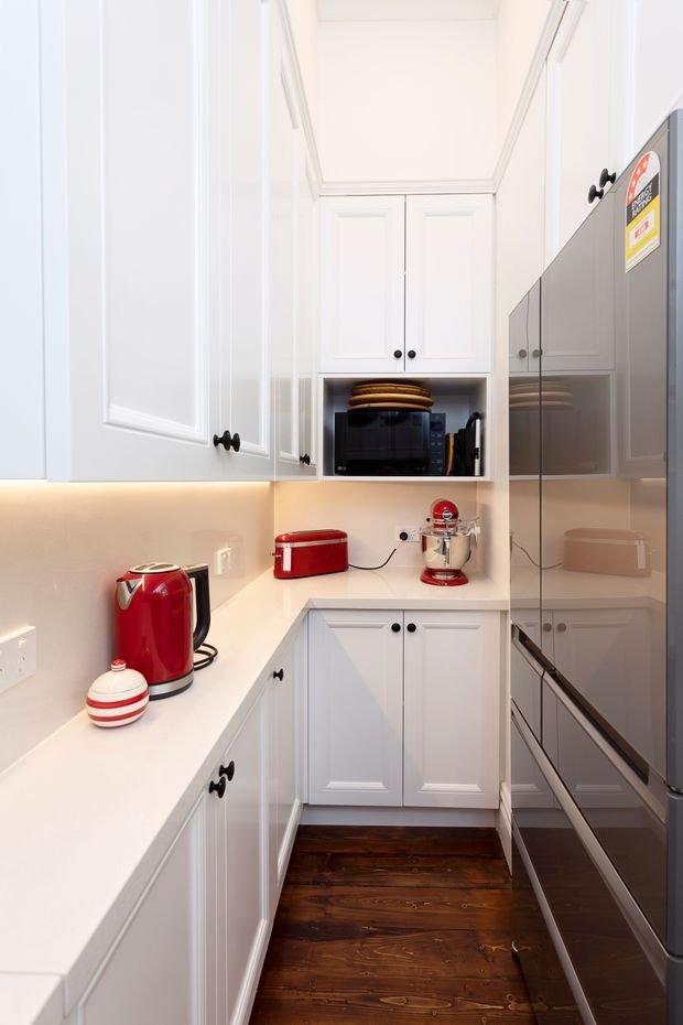 A compact, bright galley-style kitchen with white raised-panel cabinetry, dark round knobs, white benchtops, warm under-cabinet lighting, stainless-steel appliances, and a dark stained timber floor. Red countertop appliances provide bold accents, while mirrored appliance surfaces reflect the cabinetry and workspace.