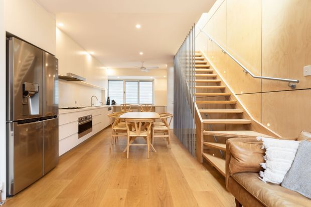 A bright open-plan kitchen and dining area with a timber staircase and partial living space. The kitchen features white flat-panel cabinetry, integrated appliances, a stainless-steel refrigerator, pale benchtops, and a window with blinds. A timber dining table and chairs sit centrally on warm timber flooring. The open-riser staircase has timber treads, a slim metal balustrade, and a vertical slatted screen, while the adjacent wall is clad in light timber panels. A leather sofa is partially visible in the foreground.