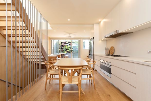 Bright open-plan kitchen and dining area with a long white built-in kitchen along the right wall, integrated stainless-steel appliances, pale timber flooring, and a round timber dining table surrounded by matching chairs. A sculptural open-riser staircase with vertical metal balusters occupies the left side, leading toward a bright glazed rear area with indoor planting visible beyond.