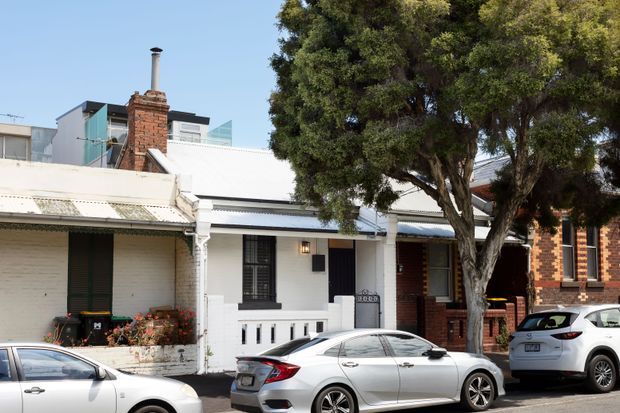 Street-level view of a row of attached urban homes, dominated by a narrow white-painted cottage-style facade with a corrugated metal verandah roof, timber detailing, dark-framed windows, and a brick chimney. Adjacent red-brick terraces and parked cars line the street, while a mature street tree partially obscures the right side. The image shows a mixed older and newer urban residential streetscape under clear daylight.