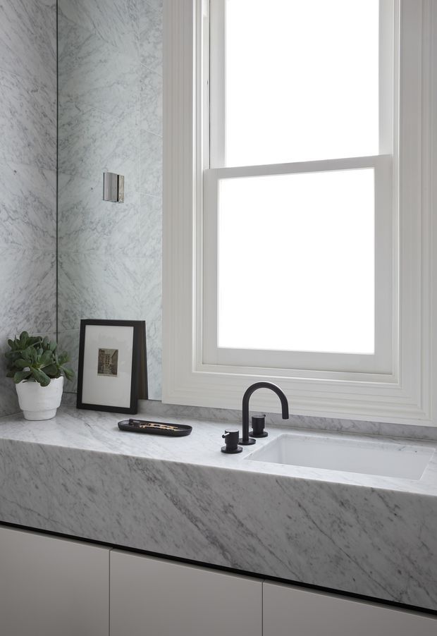 Close-up of a refined bathroom vanity beneath a large white-trimmed window. The vanity features a continuous grey-veined natural stone benchtop and integrated basin, dark curved tapware, pale flat-front cabinetry, and a marble-look wall surface. A small plant and framed artwork add restrained decoration.
