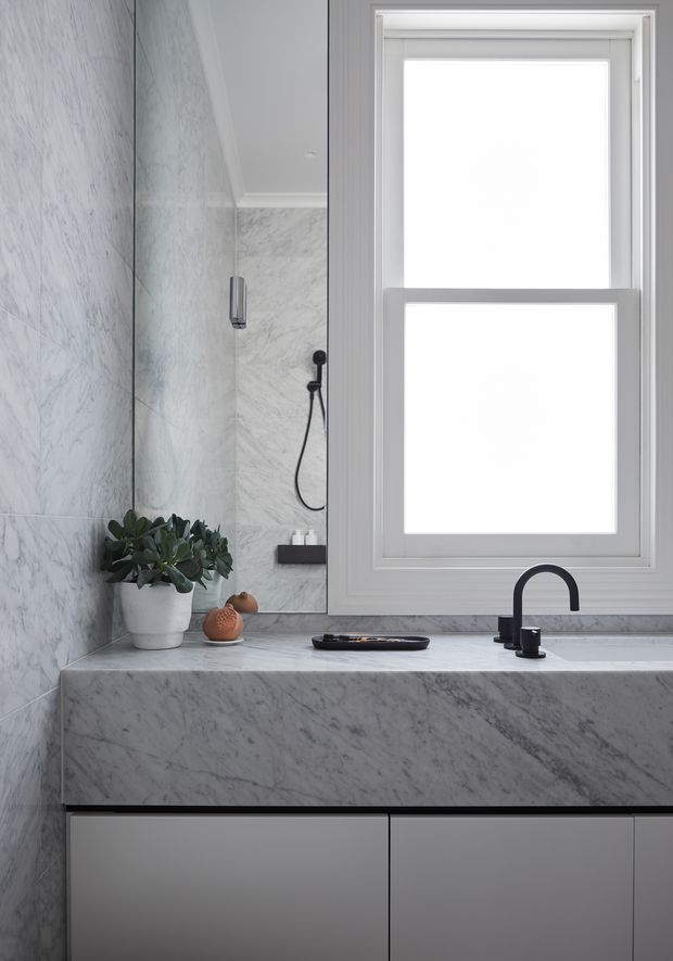 A bright, refined bathroom vanity with extensive grey-veined marble wall cladding and benchtop, sleek grey handleless cabinetry, a matte black gooseneck tap, and a large white-framed window. A glass shower enclosure is visible beside the vanity, with a small potted plant and minimal accessories on the counter.