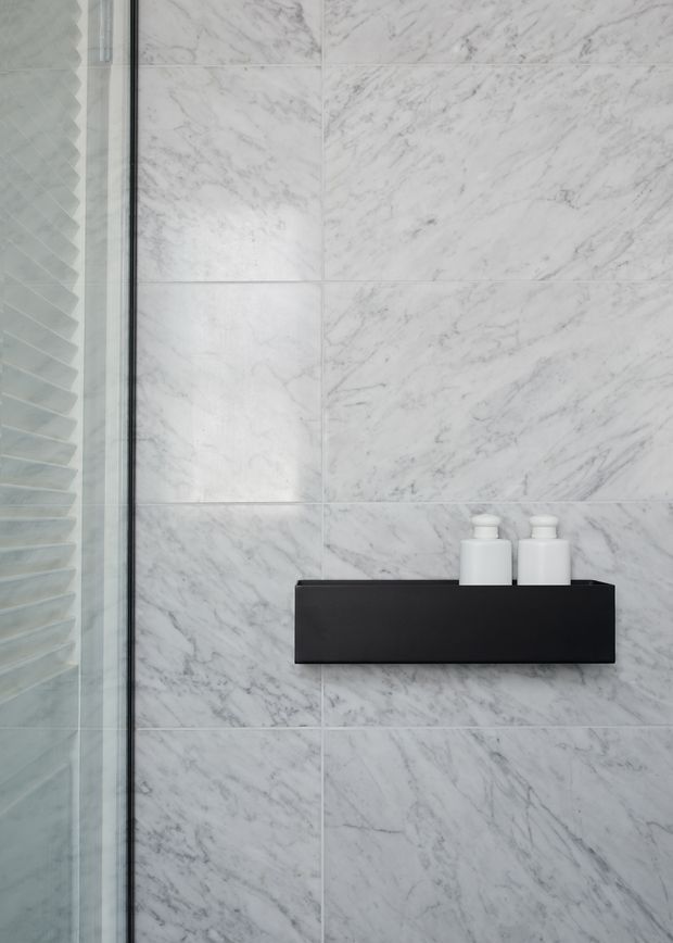 Close-up of a contemporary bathroom shower wall clad in large white marble-look tiles with grey veining. A frameless glass shower screen is visible at left, with a slim black floating shelf holding two white bottles. The composition is minimal and monochromatic.
