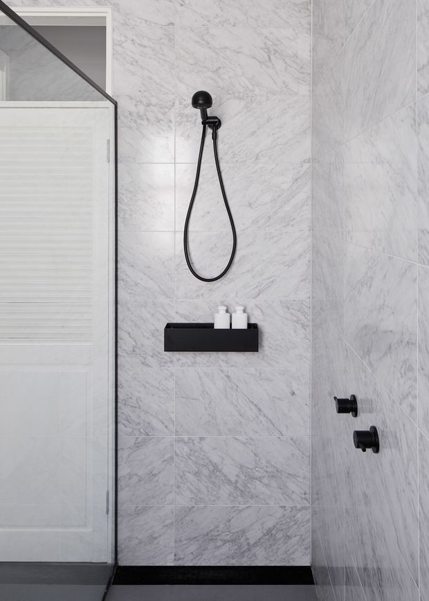 A compact contemporary shower enclosure with large white-and-grey marble-look wall tiles, matte black shower hardware, a black recessed shelf holding white bottles, a frameless glass screen, and a white paneled door. The monochromatic palette creates a clean, refined bathroom interior.