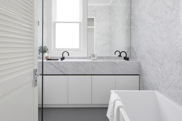 A bright, refined contemporary bathroom featuring a double vanity with pale flat-panel cabinetry, a white marble-look benchtop and matching wall finish, black tapware, a large frameless mirror, a white bathtub, and a grey floor. A louvred white door and window provide additional architectural detail.