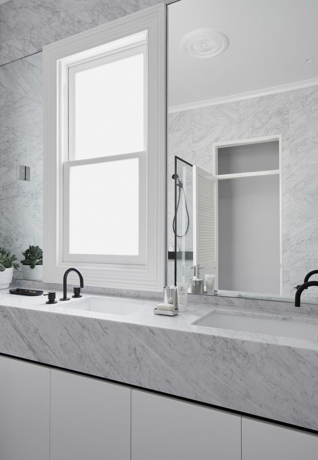 A bright, refined bathroom vanity with a large white-and-grey marble-look benchtop and backsplash, integrated double basins, matte black curved tapware, white cabinetry, a framed window, and a large mirrored wall cabinet. The shower area is visible in the mirror reflection. Decorative ceiling moulding and a ceiling medallion add a traditional architectural touch.