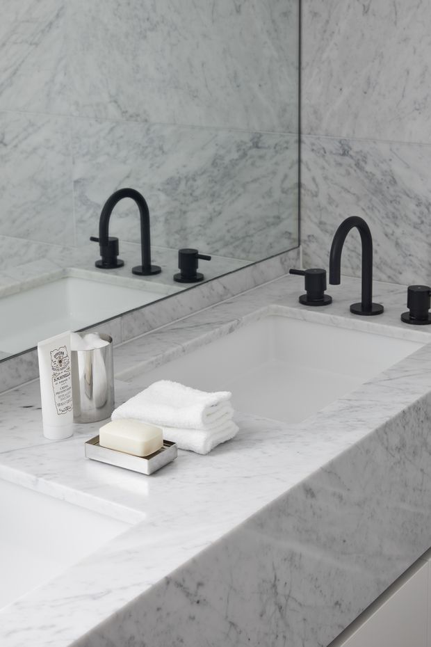 Close-up of a refined bathroom vanity with a white marble countertop and matching marble wall cladding. A rectangular undermount basin is paired with matte black arched tapware and separate controls, with a large mirror above. Folded white towels, a soap bar in a metal tray, and bathroom accessories sit on the vanity.