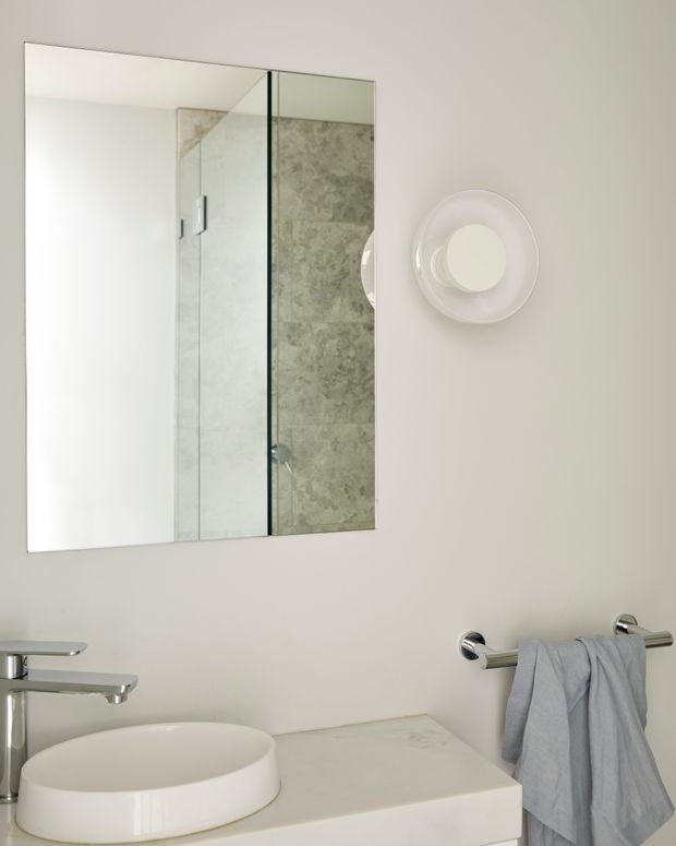 Bright, compact contemporary bathroom featuring a white vessel basin on a pale stone vanity, chrome tapware and towel rail, a frameless glass shower enclosure with grey-green stone-look wall tiles, a large mirrored cabinet, and a sculptural circular wall light.