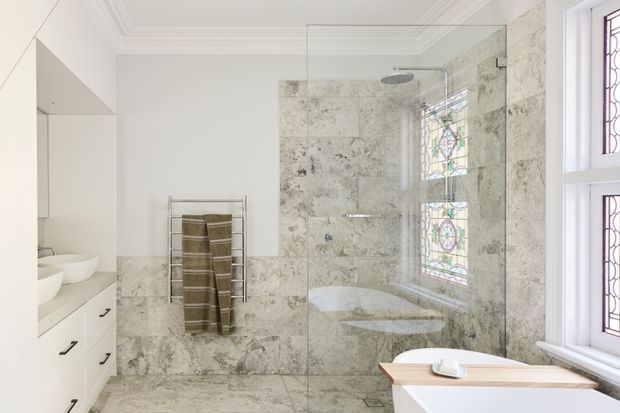 Bright, refined bathroom featuring full-height pale veined stone wall and floor finishes, a frameless glass walk-in shower with overhead rain shower, freestanding white bathtub, white vanity cabinetry with vessel basins, heated towel rail, and decorative stained-glass windows.