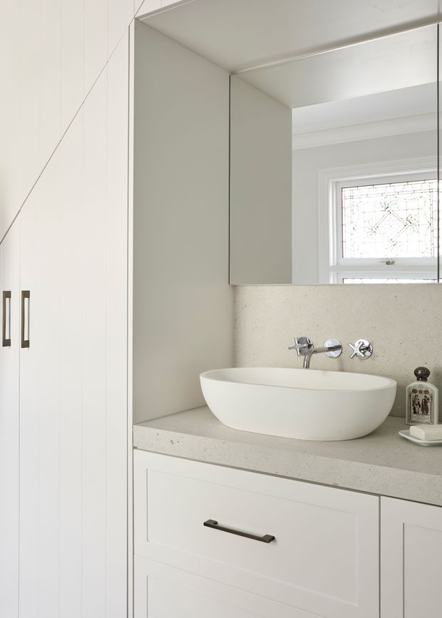 Bright, compact bathroom vanity with a white vessel basin, wall-mounted chrome tapware, pale speckled stone benchtop and matching splashback, white shaker-style cabinetry, mirrored overhead cabinet, and a decorative leadlight window. The space is clean, restrained and predominantly white with subtle warm detailing.