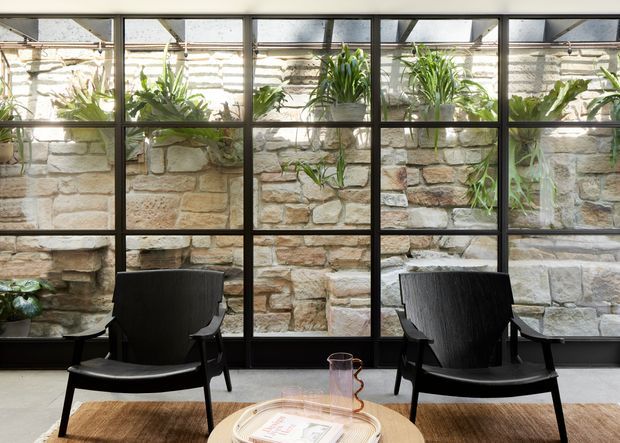 A refined interior seating area arranged in front of a full-height black-framed glazed wall. Behind the glazing is a planted stone masonry lightwell or courtyard, with abundant greenery and layered pale natural stonework. Two black lounge chairs face a small round light-wood table, creating a calm, warm, indoor-outdoor atmosphere.