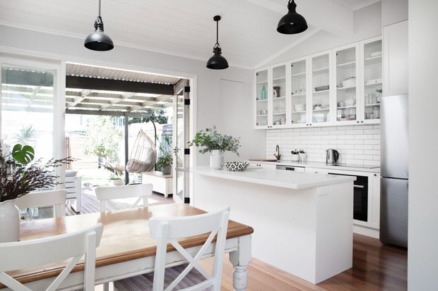 Bright open-plan kitchen and dining area with white cabinetry, glass-front upper cupboards, white subway-tile splashback, a central island, timber flooring, and a light timber dining table with white cross-back chairs. Three black pendant lights provide contrast, while large glazed doors connect the kitchen to a covered outdoor area.