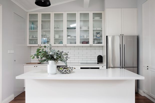 Bright, refined white kitchen with a large waterfall-style island, shaker-style cabinetry, glass-front upper cupboards, a white subway-tile splashback, and stainless-steel French-door refrigerator. Minimal decorative styling and light timber flooring create a clean contemporary look.