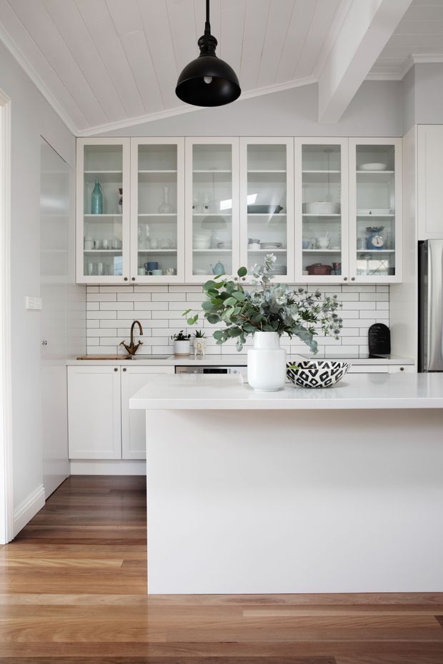 Bright, compact white kitchen with a large central island, white shaker-style cabinetry with glass-front upper doors, white subway-tile splashback, timber flooring, and a black pendant light. A sink and stainless-steel refrigerator are visible, with restrained decorative styling and greenery on the island.
