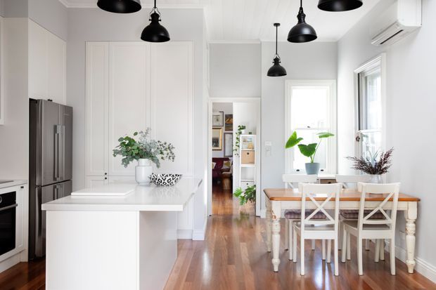 Bright open-plan kitchen and dining area with white shaker-style cabinetry, a large white island, stainless-steel refrigerator, warm timber flooring, black pendant lights, and a white dining table positioned beside tall windows. A hallway and additional living space are visible beyond.
