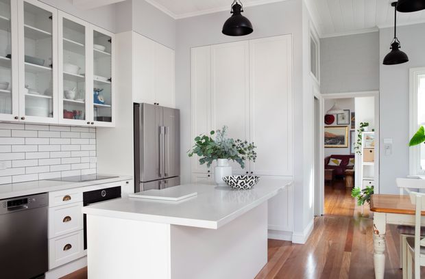 Bright, refined kitchen with white shaker-style cabinetry, glass-front upper cupboards, a white subway-tile splashback, stainless-steel refrigerator and a large white island with a pale stone benchtop. Black pendant lights contrast with the light grey walls and white decorative wall panelling. The kitchen opens toward a timber-floored hallway and an adjoining dining area.