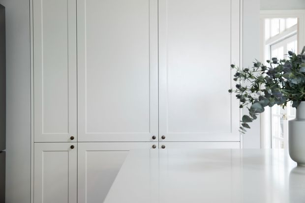 A bright, restrained interior detail showing a wall of white Shaker-style built-in cabinetry with recessed panel doors and small dark metal knobs. A white island or benchtop occupies the foreground, while a large window and a simple vase with leafy branches add natural light and greenery.