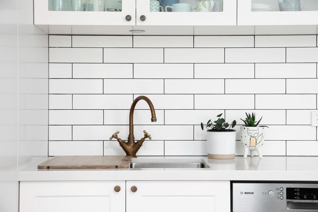 Bright, compact white kitchen with a farmhouse-style double-handle bronze tap, stainless-steel sink, pale timber benchtop section, white cabinetry with glass-fronted upper doors, and a white subway-tile splashback with dark grout. Small potted plants and ceramics add restrained decorative detail.