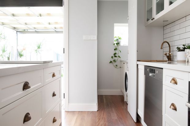 Bright, compact contemporary kitchen with white shaker-style cabinetry, brass cup pulls, white tiled splashback, stainless-steel dishwasher, and timber flooring. A laundry area is visible through the doorway, while a glazed opening leads to a light-filled outdoor or adjoining space.