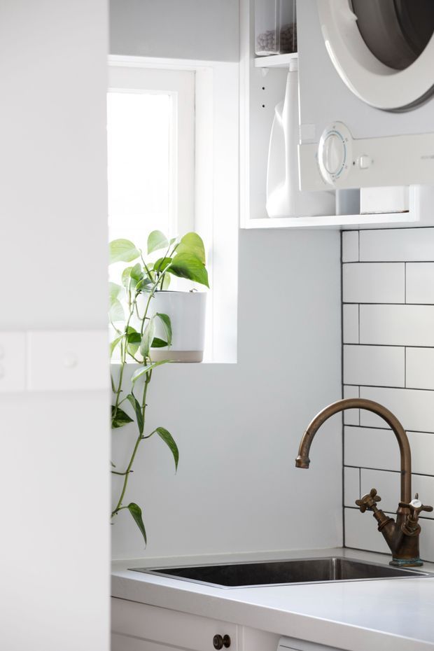 Bright, compact white kitchen corner with a stainless-steel sink, curved bronze-toned mixer tap, white subway-tile splashback, white cabinetry, open shelving, and a potted trailing plant beside a window. A white appliance is integrated into the upper cabinetry, contributing to a clean, light-filled design.