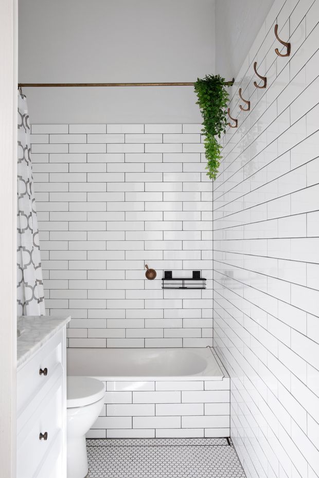 Bright compact bathroom with white subway-tiled walls, a tiled bathtub surround, a small white vanity, toilet, and grey-white hexagonal floor tiles. Copper-toned wall hooks, a black bath shelf, patterned shower curtain, and trailing greenery add contrast and detail.