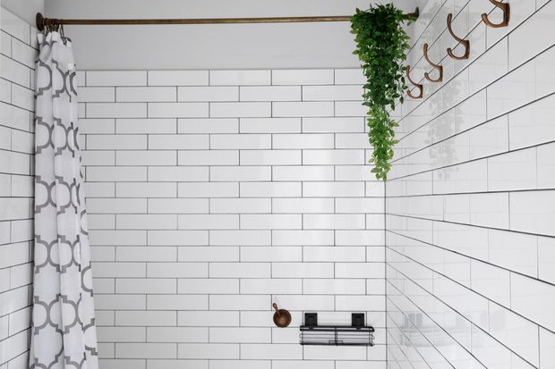 Bright bathroom or shower area clad in white horizontal subway tiles with dark grout. A patterned shower curtain hangs at left, while a brass-toned rail, hooks, shower accessories, and a hanging trailing plant add warm accents. The space appears compact, clean, and simply styled.