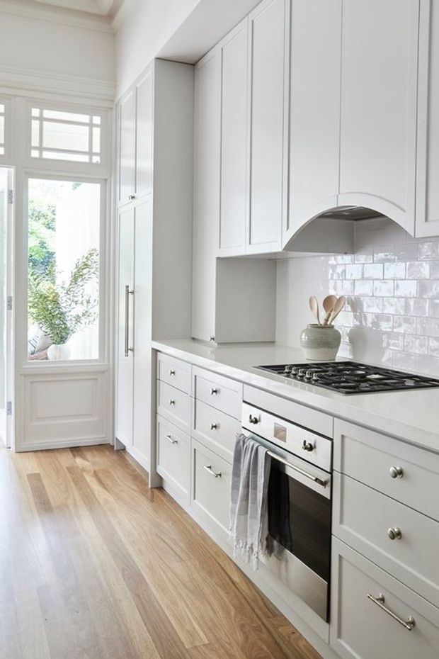 A bright, narrow galley-style kitchen with extensive white Shaker-style cabinetry, integrated tall storage, pale stone benchtops, and a glossy white handmade-look subway tile splashback. A stainless steel gas cooktop and oven sit beneath a curved feature rangehood surround. The room has a warm timber floor, traditional wall panelling and trim, and a glazed exterior door bringing in abundant natural light.