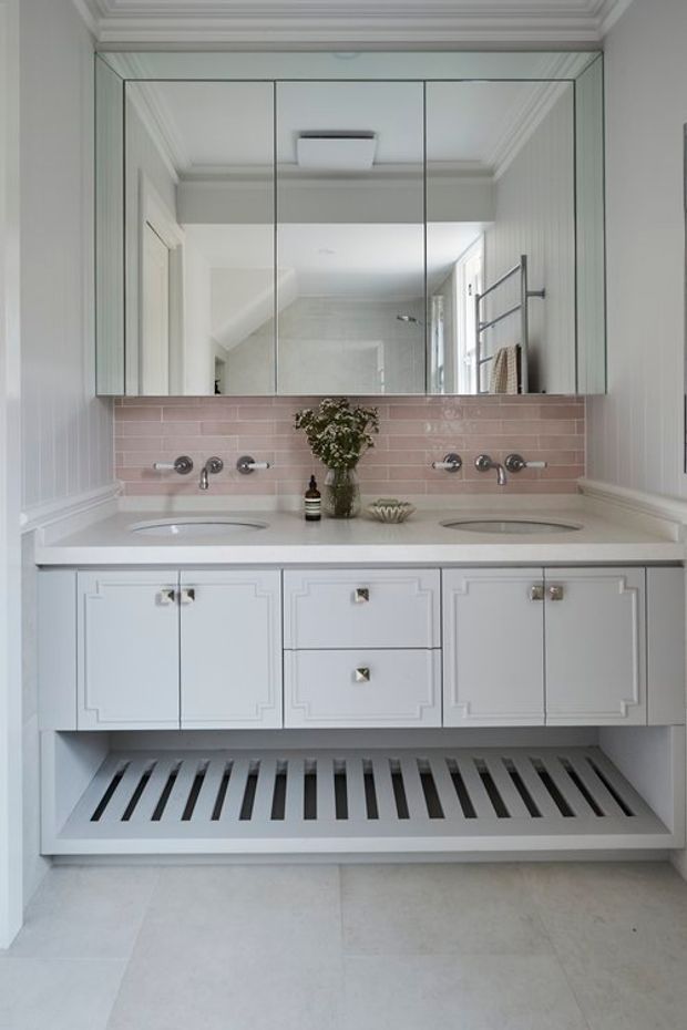 Bright, refined bathroom vanity with twin oval basins, wall-mounted chrome tapware, a large mirrored cabinet, pale pink tiled splashback, white decorative cabinetry, and a slatted open lower shelf. White panelled walls, ornate cornice detailing, and pale stone-look flooring create a classic, elegant interior.