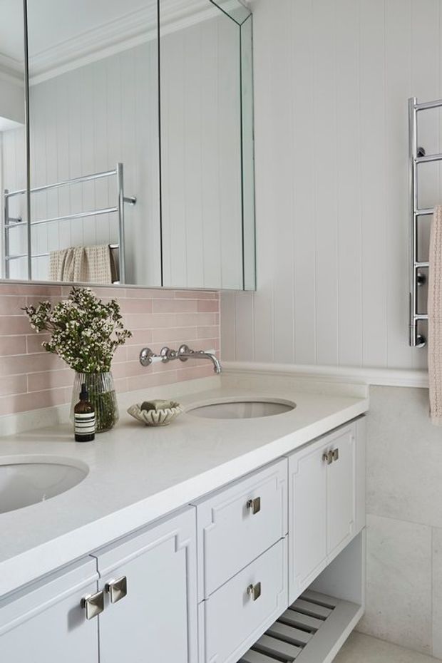 Bright, refined bathroom vanity with a long white double-sink benchtop, white paneled cabinetry, polished chrome hardware and wall-mounted tapware. The walls combine vertical white paneling with a soft blush-pink horizontal tile splashback. A large mirrored cabinet, chrome heated towel rail, and pale stone-look floor and wall surfaces complete the space.
