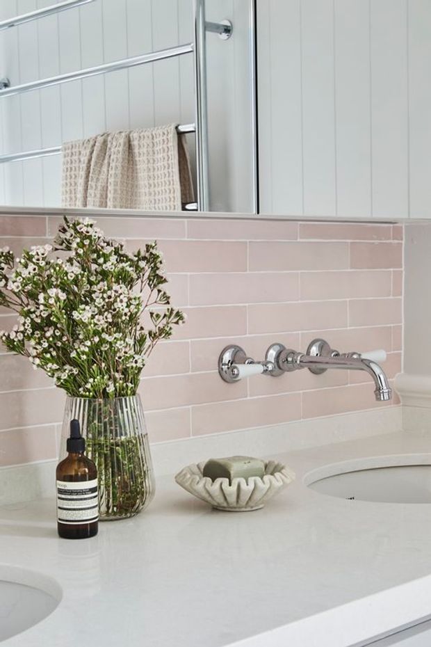 Close-up of a softly styled bathroom vanity with a pale pink horizontal tile splashback, white countertop with integrated basins, polished chrome wall-mounted tapware, a large mirror, and a towel rail with a textured neutral towel. A glass vase of flowers and soap dish add decorative detail.