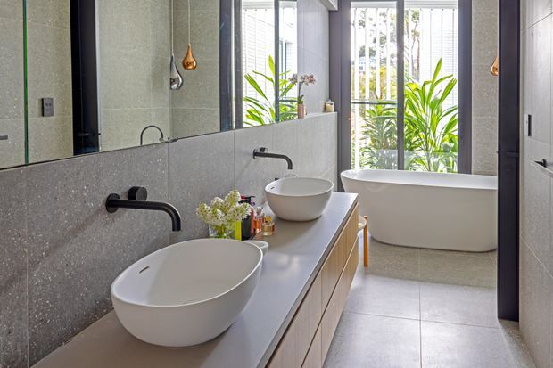 A bright contemporary bathroom with a double floating vanity, two white vessel basins, matte dark wall-mounted tapware, a large wall mirror, and a freestanding white bathtub positioned beside tall windows. Light grey stone-look wall and floor finishes create a clean, refined setting, with indoor plants visible near the glazing.