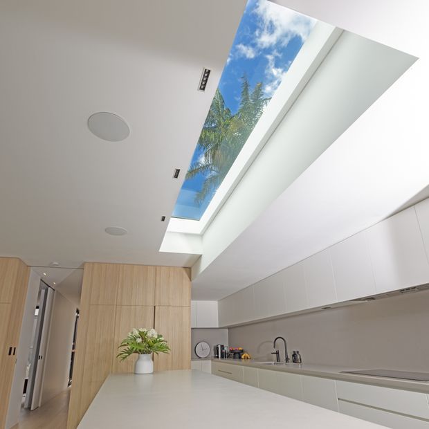 Bright contemporary kitchen with a long pale stone island, streamlined light cabinetry, integrated appliances, and a large linear skylight revealing blue sky and palm foliage. The space has a clean, minimal fit-out with light timber wall panelling, white cabinetry, recessed ceiling lights, and a muted grey backsplash.