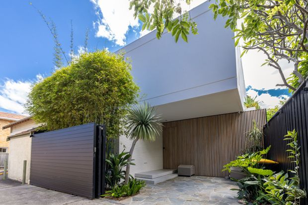 Contemporary residential facade viewed from the street, featuring a projecting upper-level white volume, dark horizontal driveway gate, vertical timber-look screening, stone-paved entry court, and lush subtropical planting. The clean geometric form contrasts with the landscaped entrance and bright blue sky.